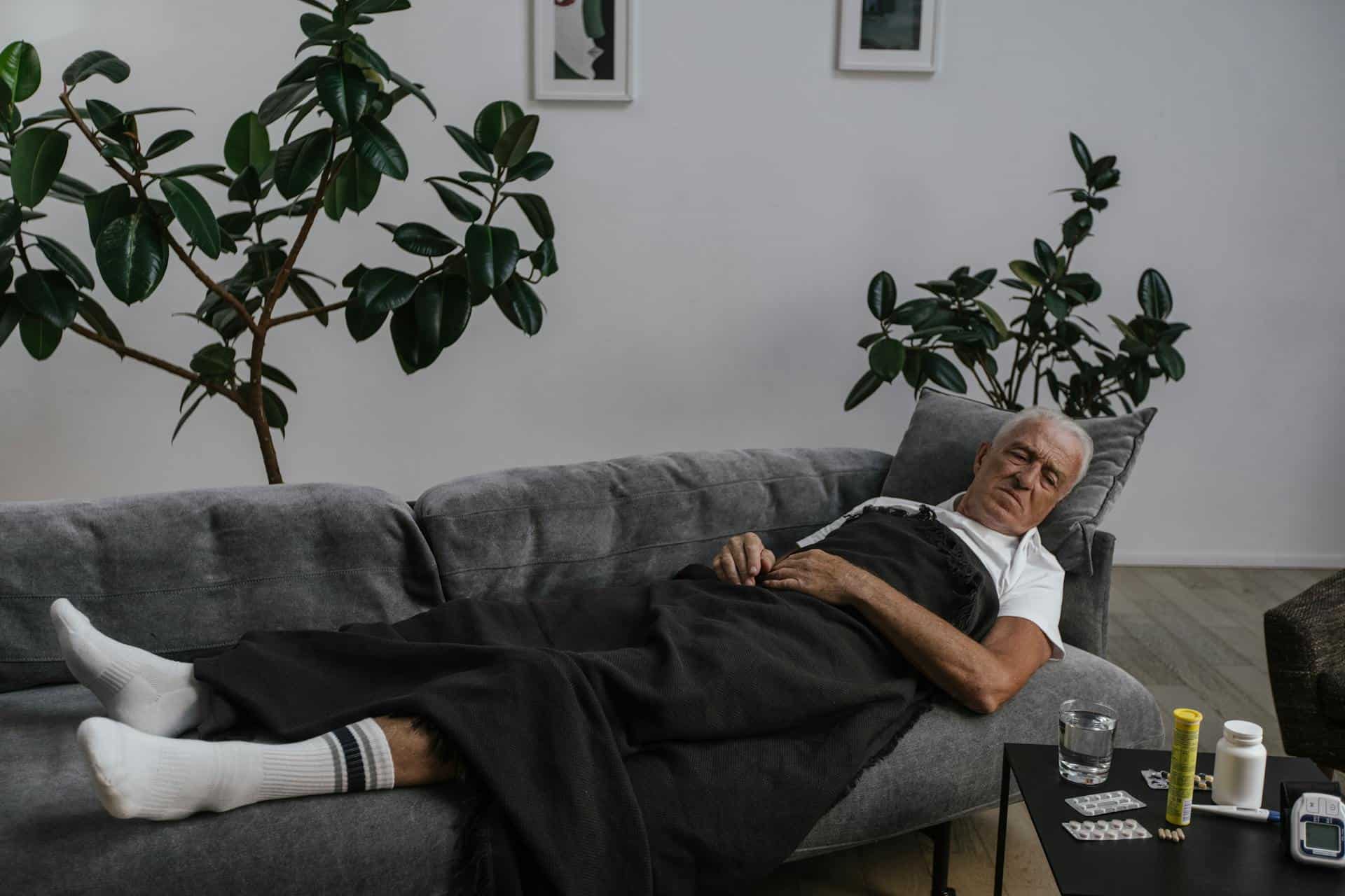 Elderly man lying on a couch under a blanket, surrounded by medication, possibly recovering from sleep-related discomfort or chronic pain.