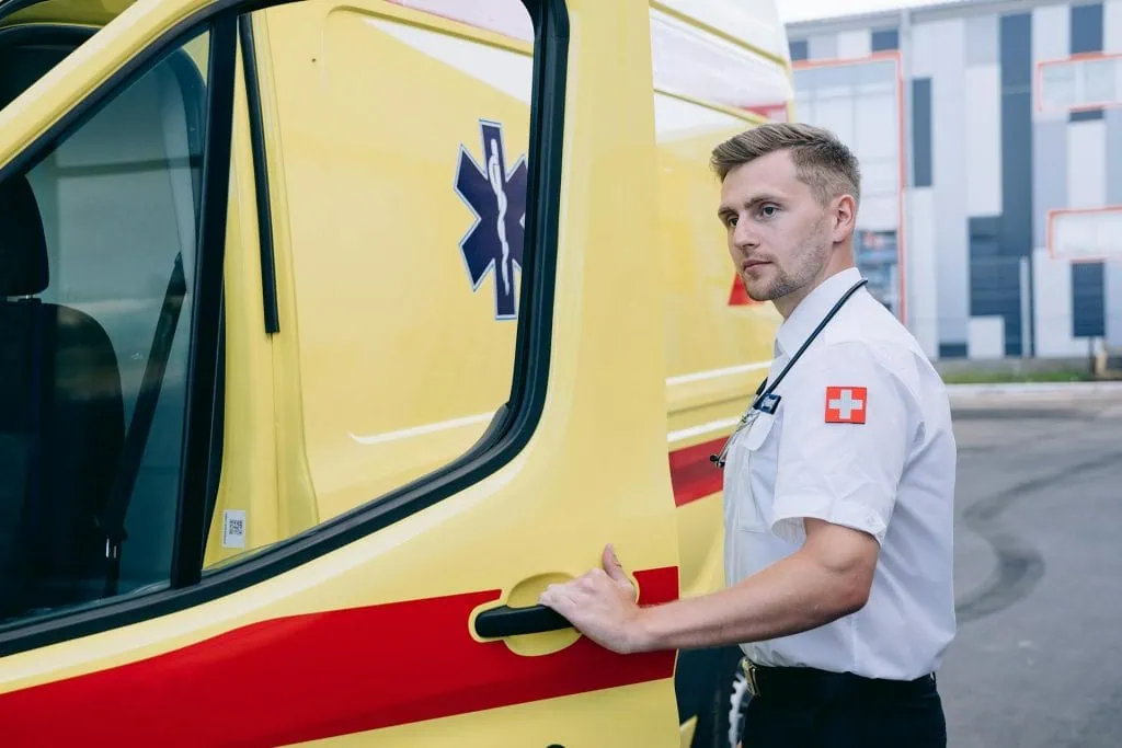 Male paramedic in uniform with a stethoscope standing beside a yellow ambulance, preparing to open the door.