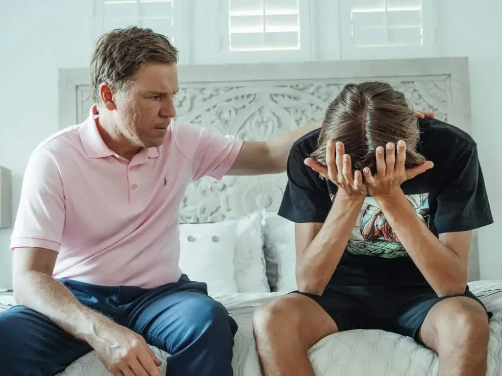 Father comforting his teenage son who appears overwhelmed and stressed, sitting on a bed in a bright room.