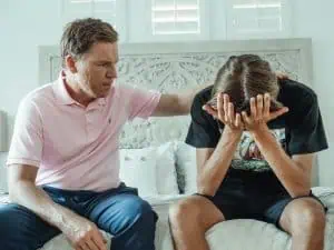 Father comforting his teenage son who appears overwhelmed and stressed, sitting on a bed in a bright room.