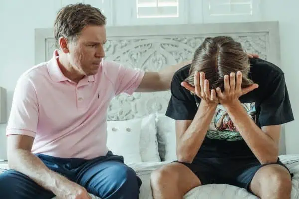 Father comforting his teenage son who appears overwhelmed and stressed, sitting on a bed in a bright room.