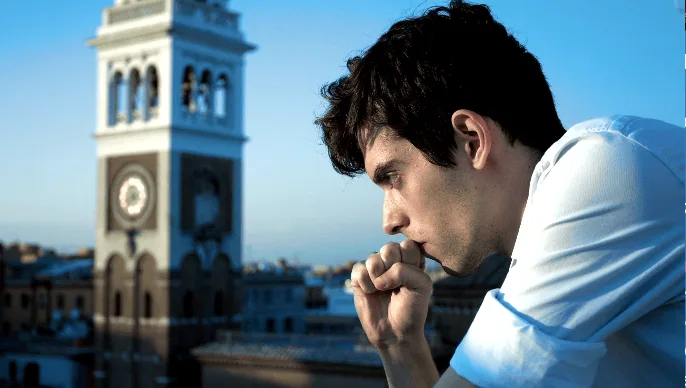 A young man in a white shirt standing on a rooftop, deep in thought with his hand near his mouth. The background features a cityscape with a clock tower, adding to the contemplative mood.