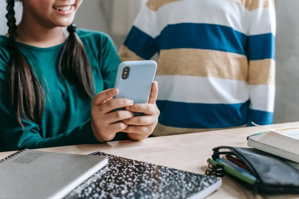 Smiling young girl using a smartphone while sitting at a desk with books and school supplies.