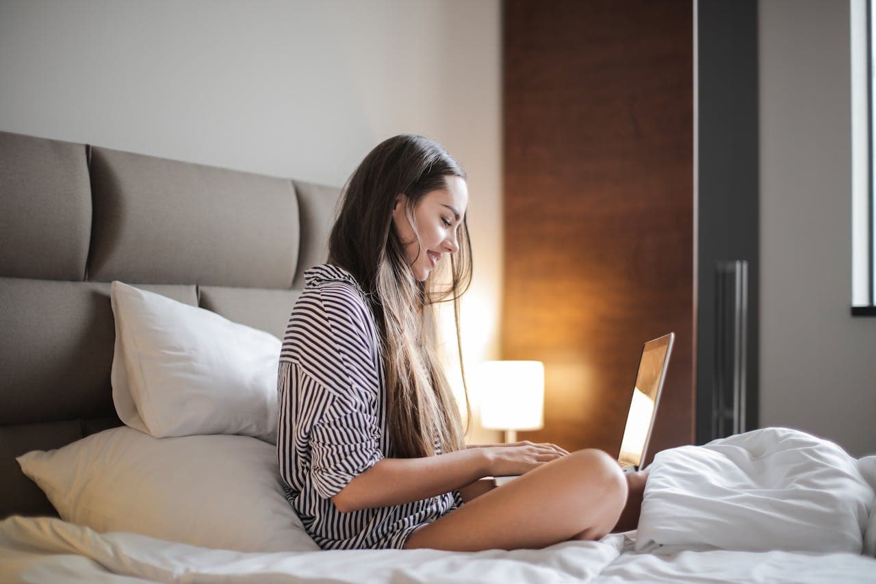sleep_Hypno relaxation at home: A young woman sitting on a bed, using a laptop with a calm and focused expression, creating a peaceful and cozy atmosphere.