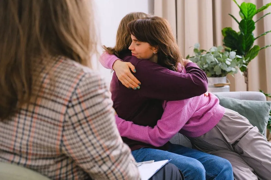 hypnotherapy_Two women embracing during a therapy session while a therapist observes and takes notes in a supportive counseling environment.