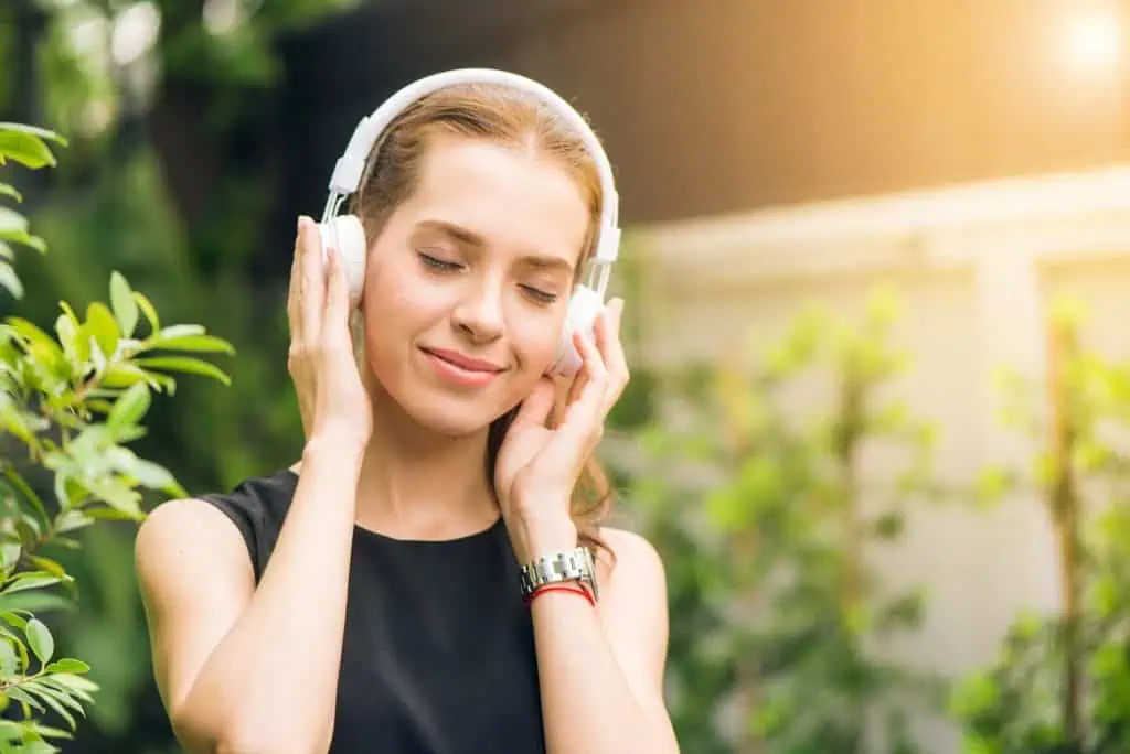 A woman enjoying music with white headphones, eyes closed and smiling, standing outdoors surrounded by greenery with sunlight in the background.