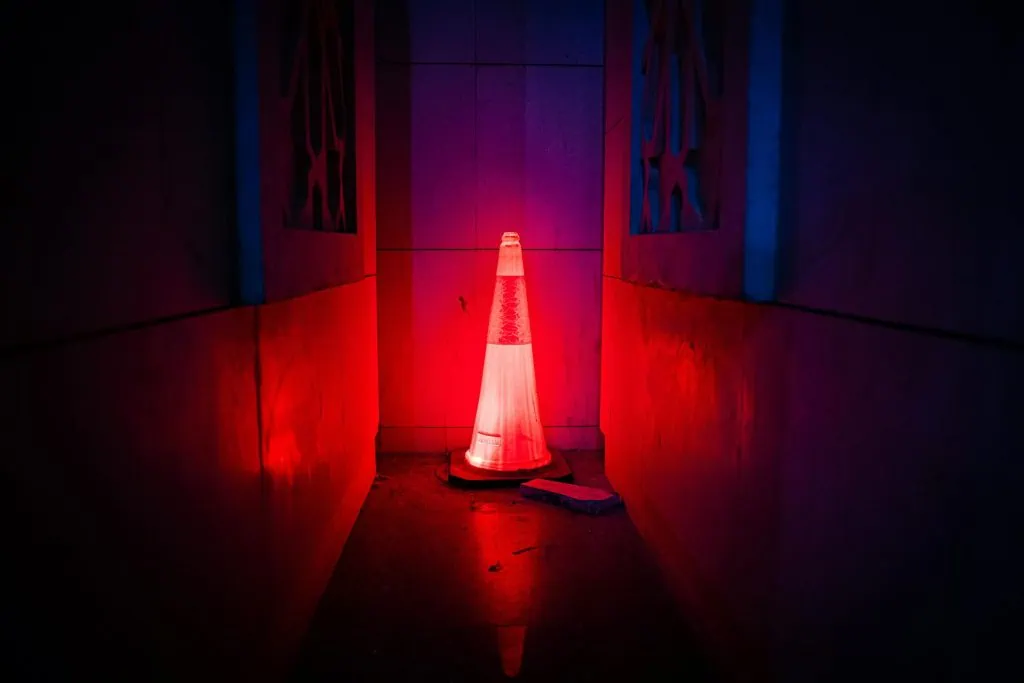 A traffic cone glowing under intense red lighting, placed in a narrow, dimly lit corridor with a moody and mysterious atmosphere. The surrounding walls reflect the red and blue hues, adding to the dramatic effect.