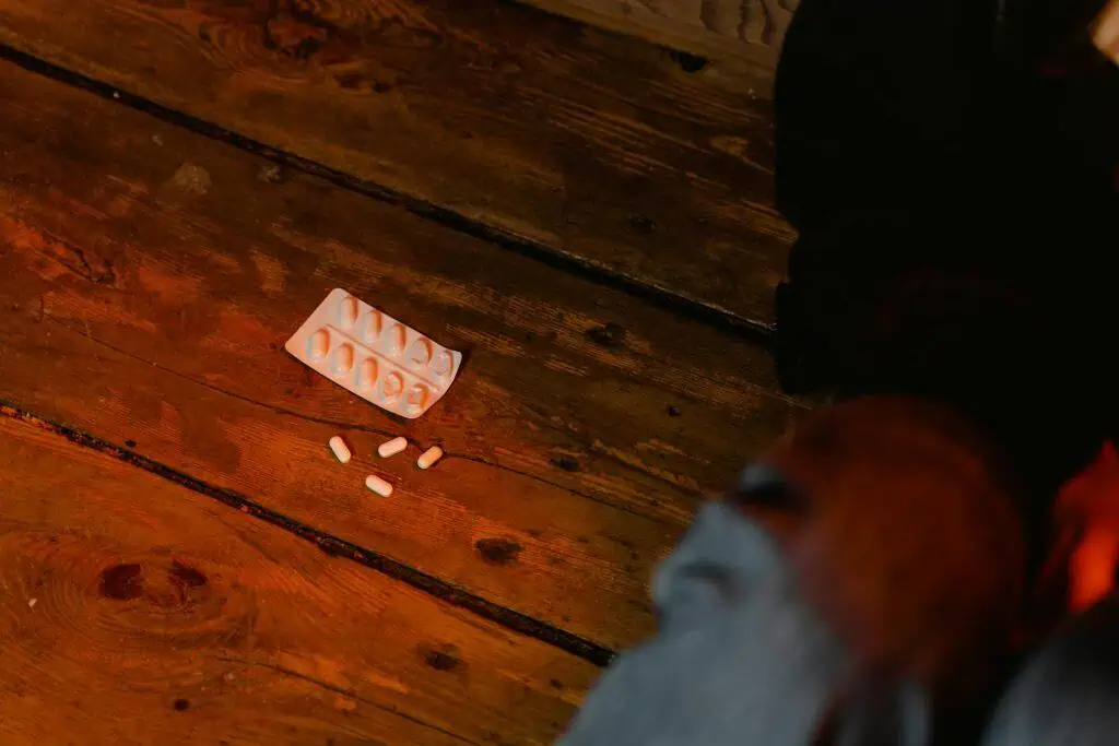 A blister pack and scattered white capsules lie on a wooden floor near a person's legs, suggesting a moment of distress or crisis.