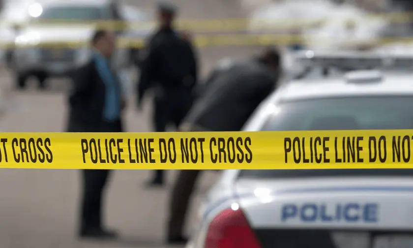 A yellow "POLICE LINE DO NOT CROSS" tape stretches across a crime scene, blocking access. In the blurred background, police officers and investigators are seen near a patrol car, assessing the situation.