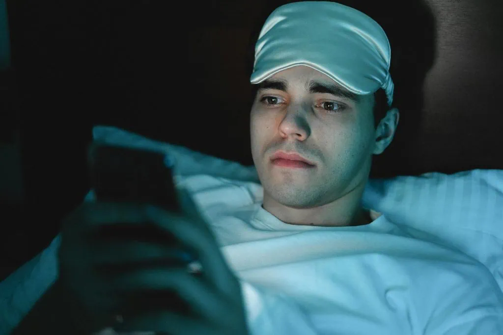 A person lies in bed at night illuminated by the glow of a smartphone screen, illustrating poor sleep hygiene habits such as screen use before bedtime that can disrupt restful sleep.