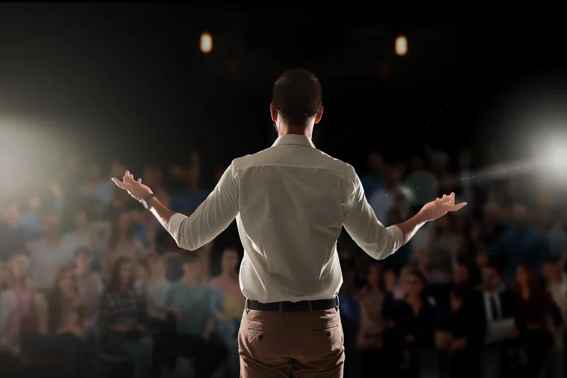 A man in a white shirt speaking to a large audience on stage under bright lights.