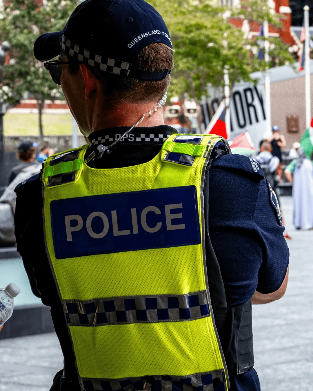 Back view of a Queensland police officer wearing a high-visibility vest labeled "POLICE" while monitoring a public event or crowd.