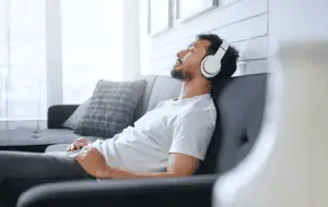 A relaxed man reclining on a couch with his eyes closed, wearing white headphones, enjoying music or an audio session in a bright and cozy living space.