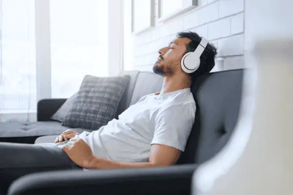 A relaxed man reclining on a couch with his eyes closed, wearing white headphones, enjoying music or an audio session in a bright and cozy living space.