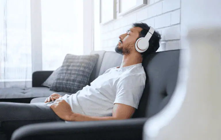 anxiety_A relaxed man reclining on a couch with his eyes closed, wearing white headphones, enjoying music or an audio session in a bright and cozy living space.