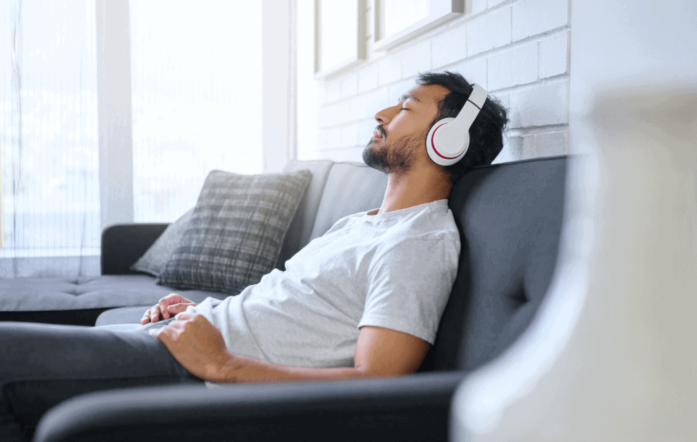 anxiety_A relaxed man reclining on a couch with his eyes closed, wearing white headphones, enjoying music or an audio session in a bright and cozy living space.