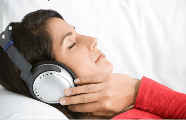 A woman in a red long-sleeve shirt lying on a white surface with her eyes closed, listening to music through large over-ear headphones. She appears calm and relaxed, enjoying a moment of peace.