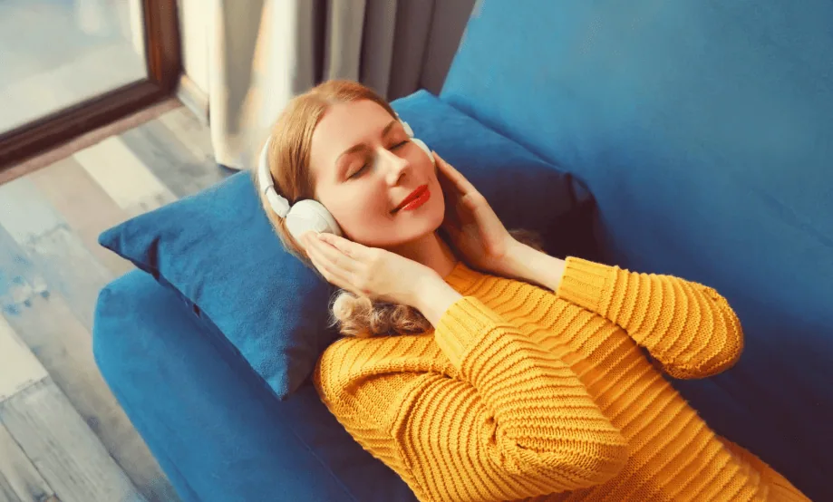 A woman in a mustard yellow sweater lying on a blue couch, listening to music through white over-ear headphones with a peaceful expression.