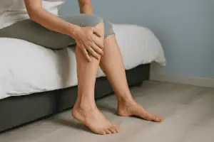 A woman sitting on the edge of a bed gently holding her leg, depicting discomfort from restless leg syndrome in a calm bedroom setting.