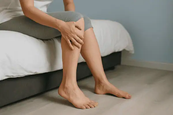 A woman sitting on the edge of a bed gently holding her leg, depicting discomfort from restless leg syndrome in a calm bedroom setting.
