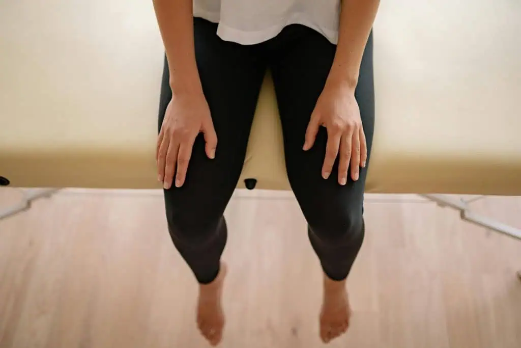 A person sitting barefoot on a therapy bed with hands resting on their thighs, representing a calm and focused moment during a consultation for Restless Legs Syndrome.