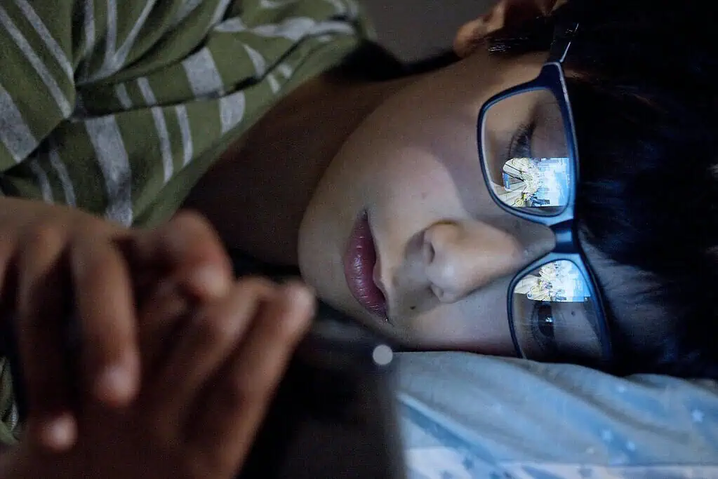 Close-up of a boy lying in bed using a tablet, with the screen’s reflection visible in his glasses, showing the link between screens and ADHD.