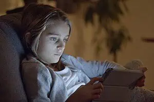 Child with focused expression using a tablet in a dimly lit room, illustrating the link between screens and ADHD.