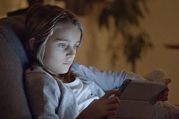 Child with focused expression using a tablet in a dimly lit room, illustrating the link between screens and ADHD.