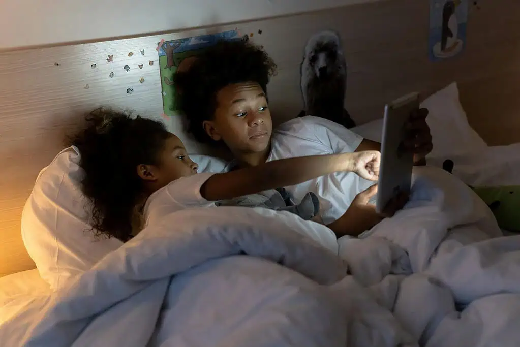Two children lying in bed using a tablet at night, showing how screens and ADHD can affect sleep and focus.