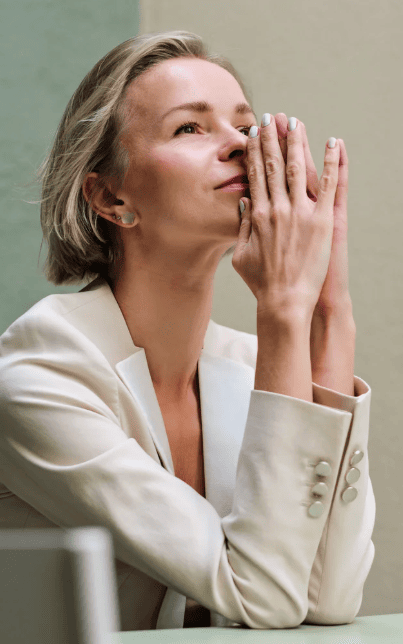 Woman in a light-colored blazer resting her hands on her face, looking upward with a peaceful and reflective expression.