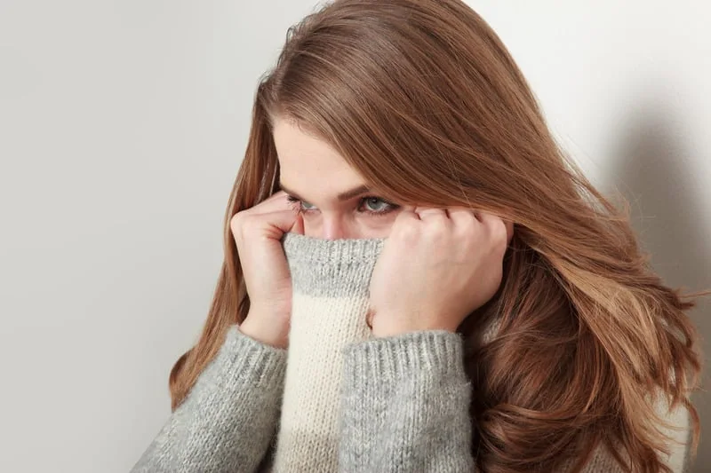 A woman with long hair hiding part of her face with a cozy sweater, looking shy or nervous.