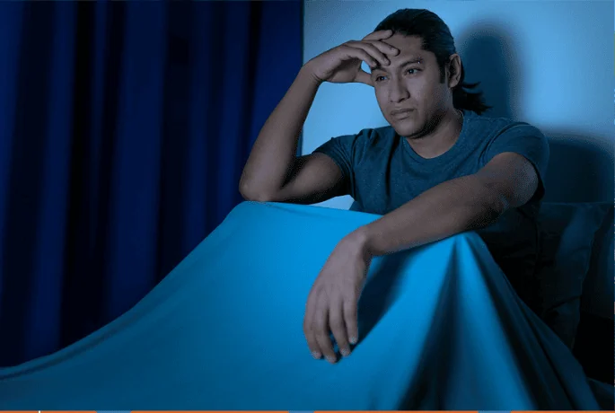 A man sitting up in bed at night, looking distressed with his hand on his forehead. The blue lighting and dark curtains suggest insomnia or deep thoughts.