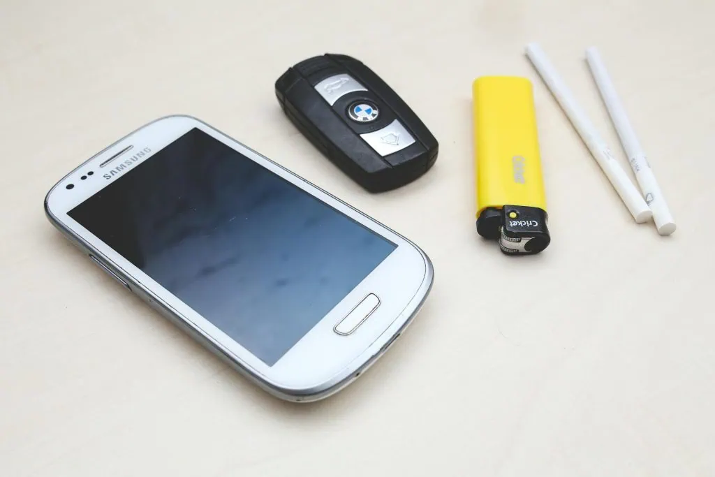 Flat lay of a Samsung smartphone, BMW car key, yellow lighter, and two white cigarette-style objects on a light wooden surface.