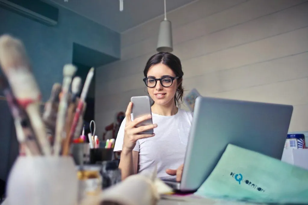 Young woman wearing glasses using a smartphone while working on a laptop in a creative workspace.