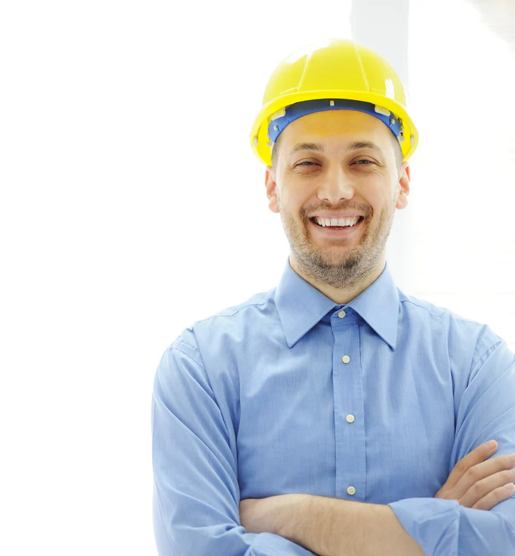 A smiling man wearing a yellow construction helmet and a light blue button-up shirt stands confidently with his arms crossed, conveying a sense of professionalism and satisfaction in his work.