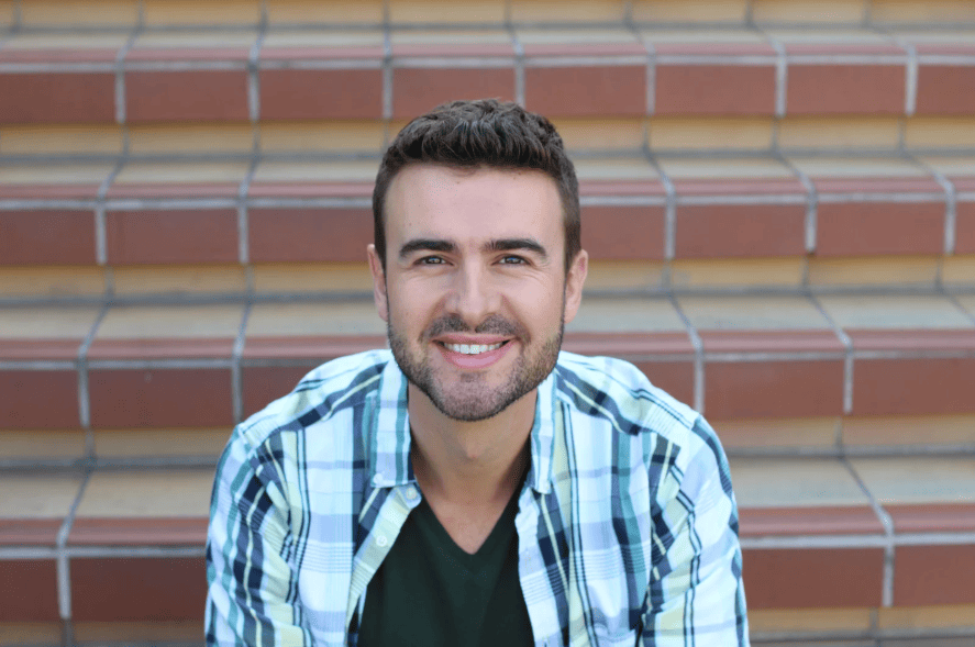 Smiling young man wearing a plaid shirt sitting on outdoor steps.