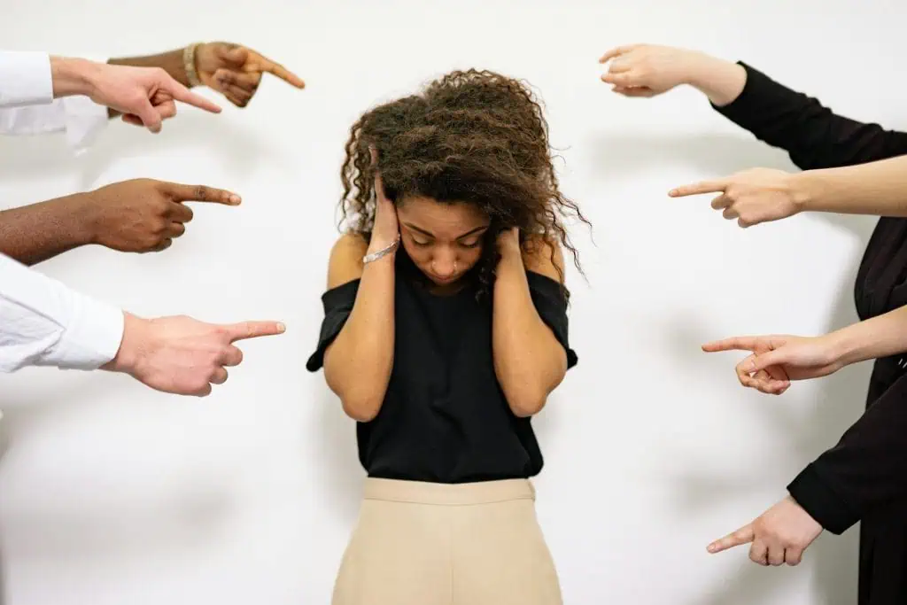 A distressed woman covering her ears while multiple hands point at her, symbolizing social anxiety and fear of judgment.
