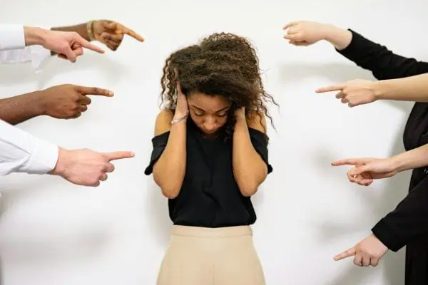 A distressed woman covering her ears while multiple hands point at her, symbolizing social anxiety and fear of judgment.