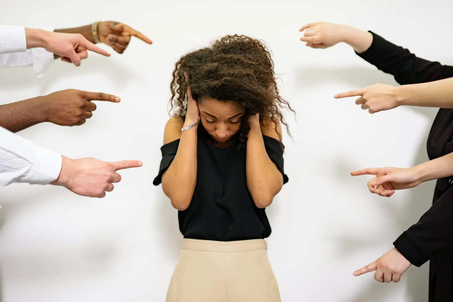 A distressed woman covering her ears while multiple hands point at her, symbolizing social anxiety and fear of judgment.
