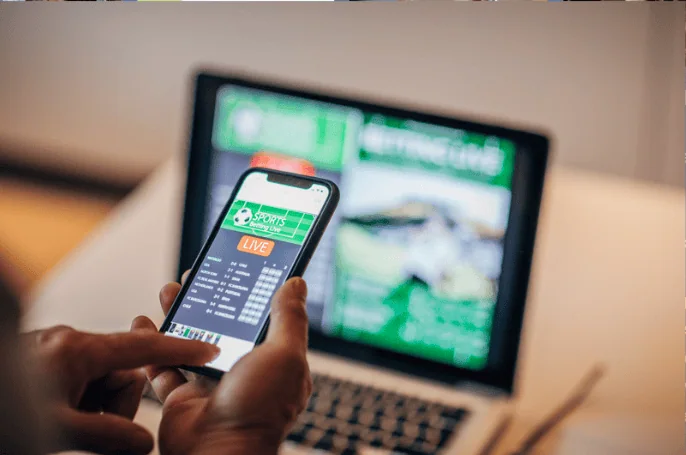 A person using a smartphone to place a live sports bet while a betting website is open on a laptop screen in the background. The focus is on the gambling activity.