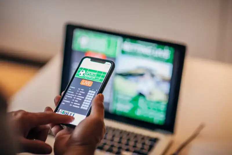 A person holds a smartphone displaying a live sports betting app while sitting in front of a laptop showing a blurred betting website, highlighting the digital nature of online sports gambling.