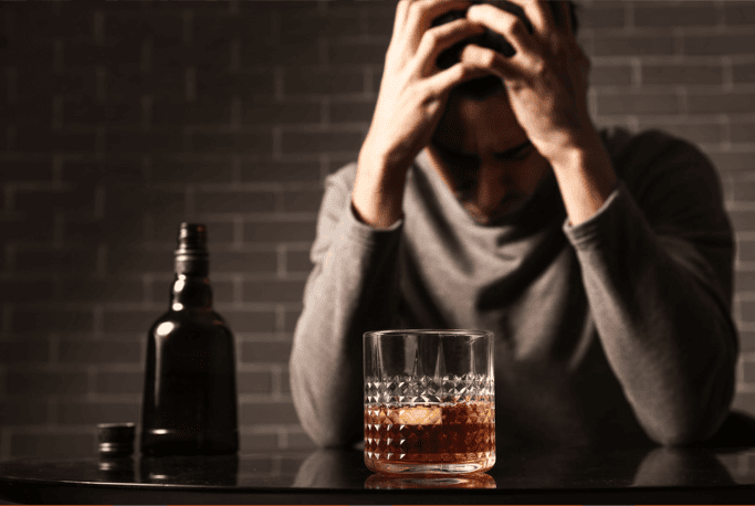 A man in a gray sweater sitting at a table, holding his head in distress. In front of him is a glass of whiskey and a bottle, suggesting he is struggling with alcohol consumption and emotional stress.