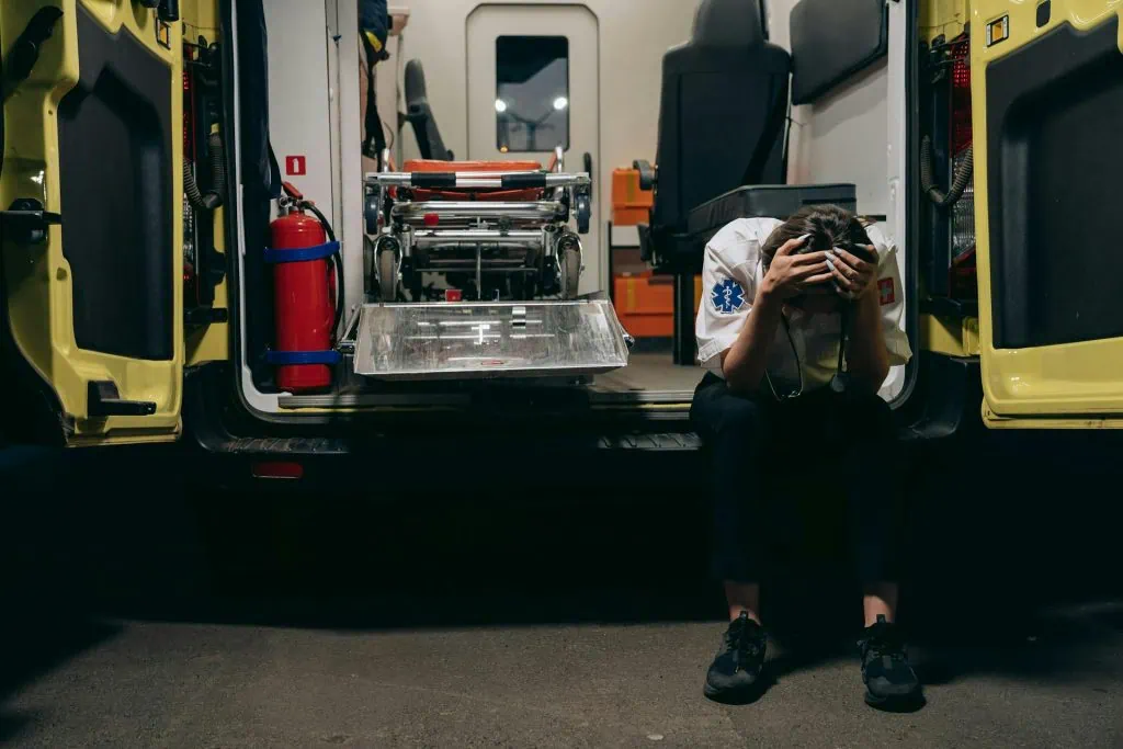 Stressed paramedic sitting on the back of an ambulance with hands on head, exhausted from long shifts
