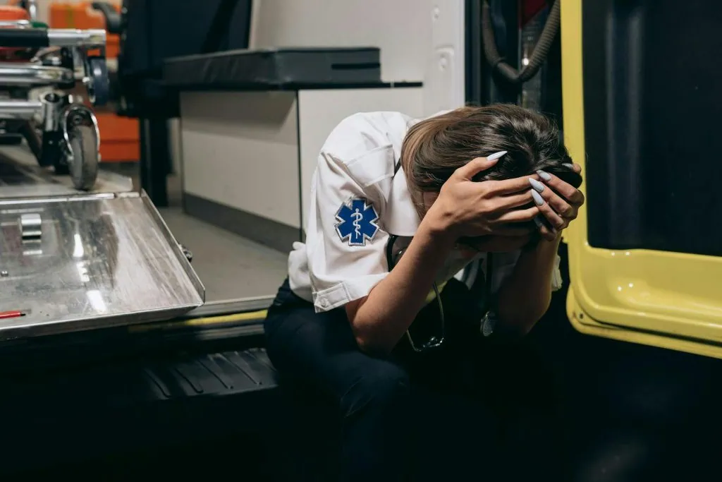 A paramedic in uniform sits with their head in their hands, showing signs of emotional exhaustion or burnout, highlighting the mental health challenges faced by emergency responders.