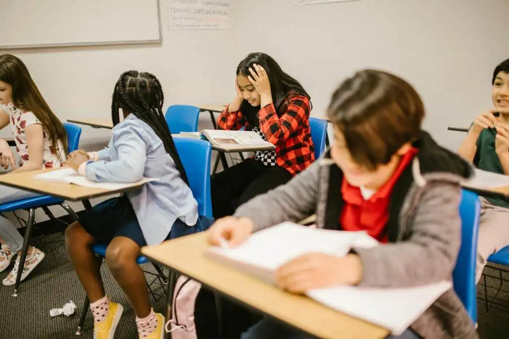 Young student holding her head in frustration while studying in a classroom, surrounded by other classmates focused on their work.