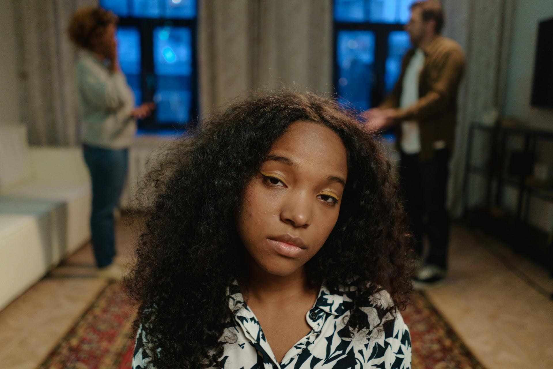 A teenage girl with curly hair looks at the camera with a weary expression while two adults argue in the room behind her