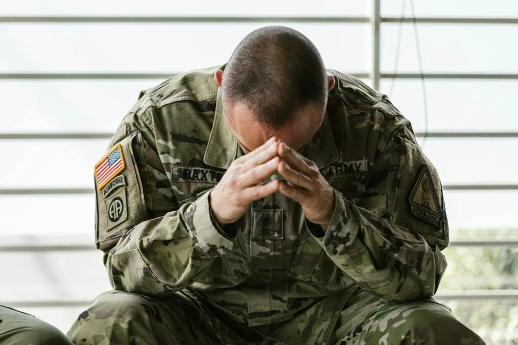 U.S. Army soldier in uniform sitting with head bowed and hands clasped, showing signs of stress or deep thought indoors.