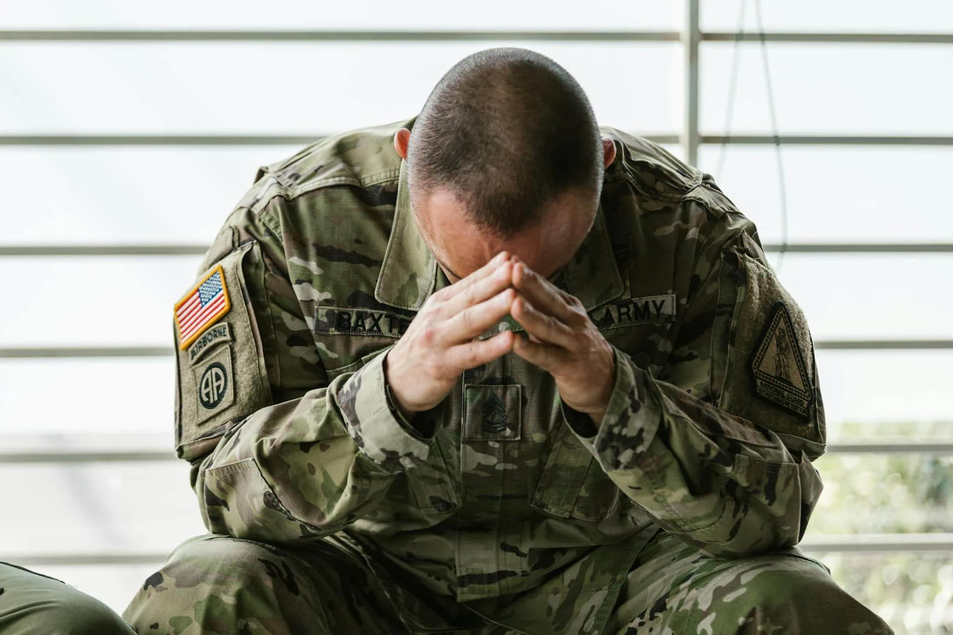 U.S. Army soldier in uniform sitting with head bowed and hands clasped, showing signs of stress or deep thought indoors.