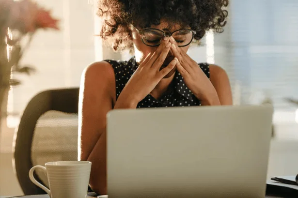 A woman with curly hair and glasses sitting at a desk, rubbing her eyes in frustration while working on a laptop, showing signs of stress or exhaustion.
