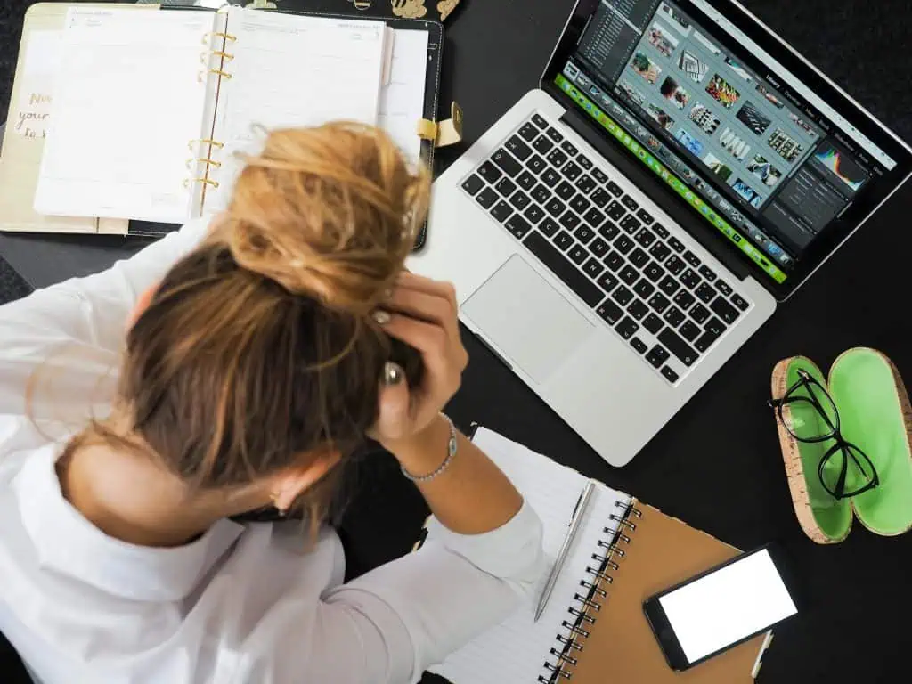 Overhead view of a woman sitting at a cluttered desk with her hands on her head, looking stressed while surrounded by a laptop, planner, notebook, smartphone, and glasses.